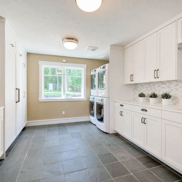 Przestronna pralnia z białymi szafkami, nowoczesnymi urządzeniami i jasno oświetlonym oknem.
