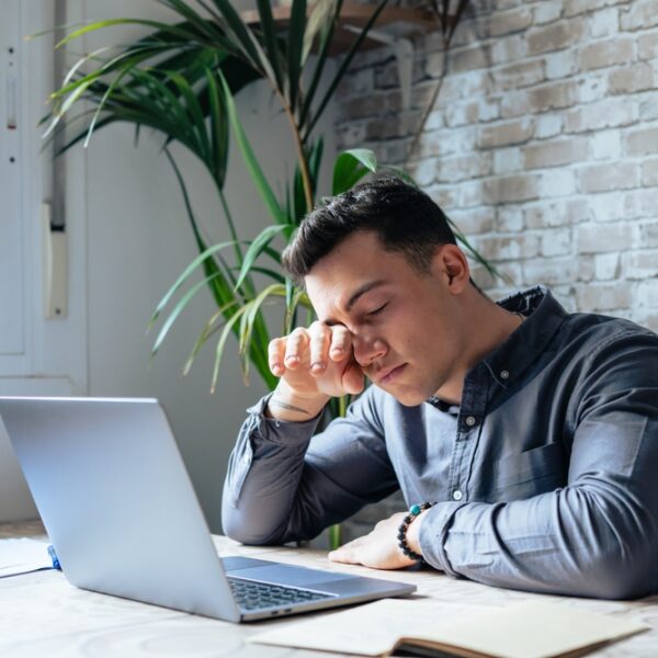 Mężczyzna w szarej koszuli siedzi przy biurku, trzymając się za czoło, obok laptop i notatki. W tle rośnie roślina.