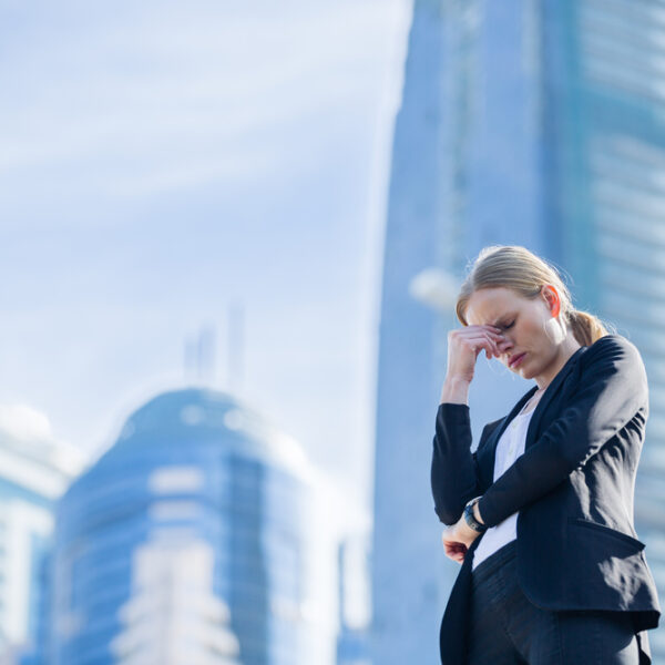 Kobieta w garniturze stoi na tle nowoczesnych budynków, trzymając się za czoło, wygląda na zmartwioną.