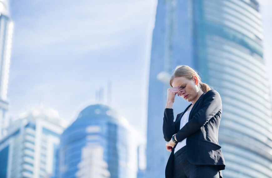 Kobieta w garniturze stoi na tle nowoczesnych budynków, trzymając się za czoło, wygląda na zmartwioną.